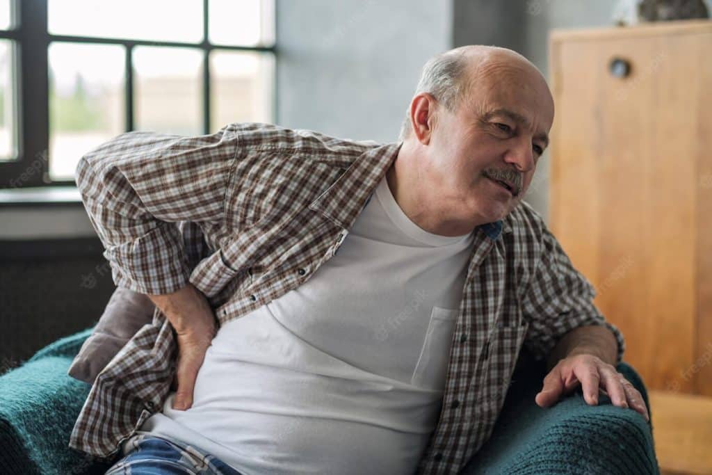 Oudere man met geruite blouse en wit T-shirt zit voorovergebogen en drukt zijn hand tegen de flank ter hoogte van de nierstreek, wat kan wijzen op flankpijn.