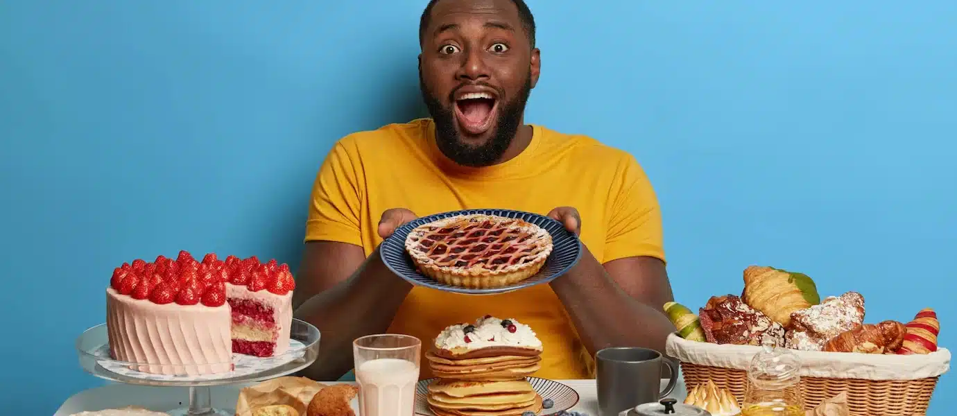 Een man met een donkere huidskleur en een baard, die een geel T-shirt draagt. Hij heeft een open mond en kijkt verrast of blij, terwijl hij een kom met eten omhoog houdt. Op de tafel voor de man ligt een grote hoeveelheid gevarieerd eten, waaronder een stapel pannenkoeken, een glas melk, en diverse andere lekkernijen die eruitzien als junkfood of ontbijtgerechten.
