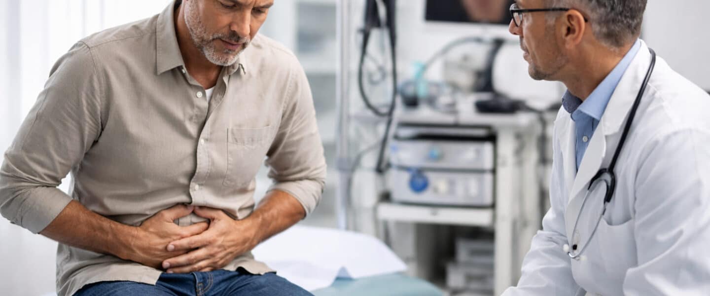 Man met buikpijn bij specialist in spreekkamer; arts luistert terwijl patiënt zijn buik vasthoudt