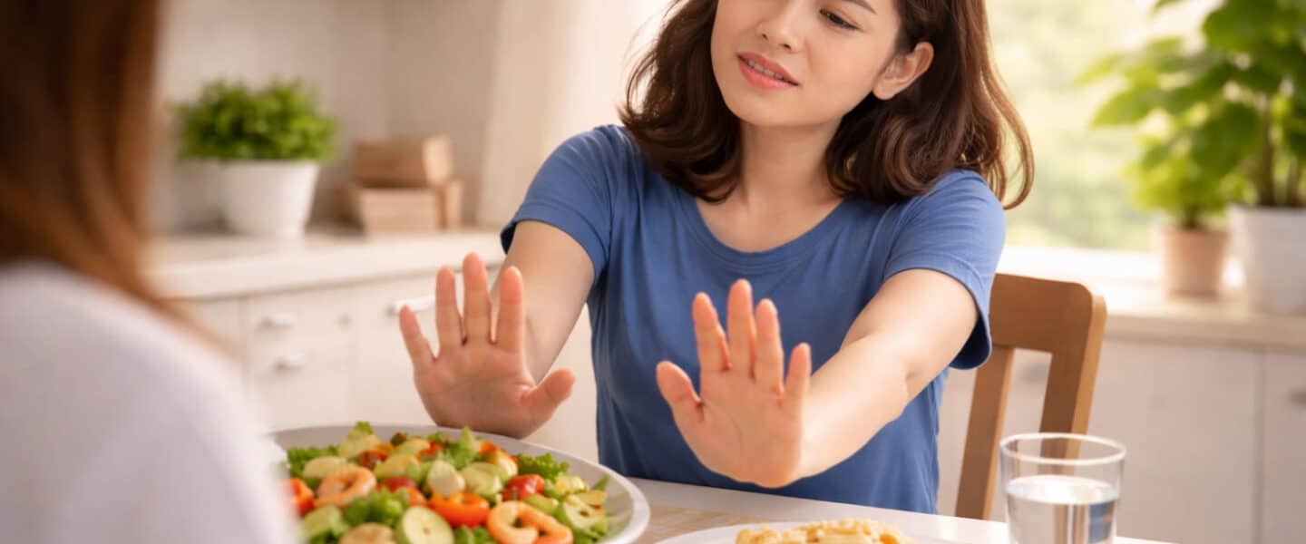 Realistische foto van een jonge vrouw die met afwerend gebaar een bord salade wegduwt terwijl zij alleen eenvoudige voedingsmiddelen op tafel accepteert, als beeld van sterke voedselvermijding bij ARFID.