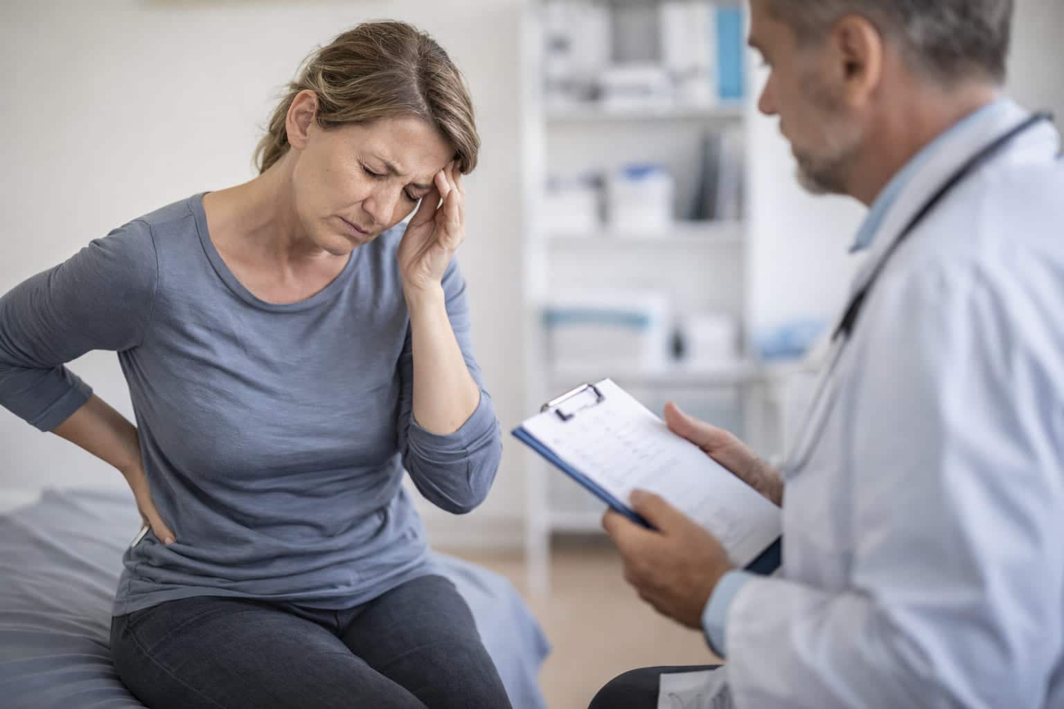 Vrouw van middelbare leeftijd zit tijdens een consult bij de huisarts met hand tegen het hoofd en pijn in de onderrug, een beeld van lichamelijke klachten zonder duidelijke medische oorzaak.