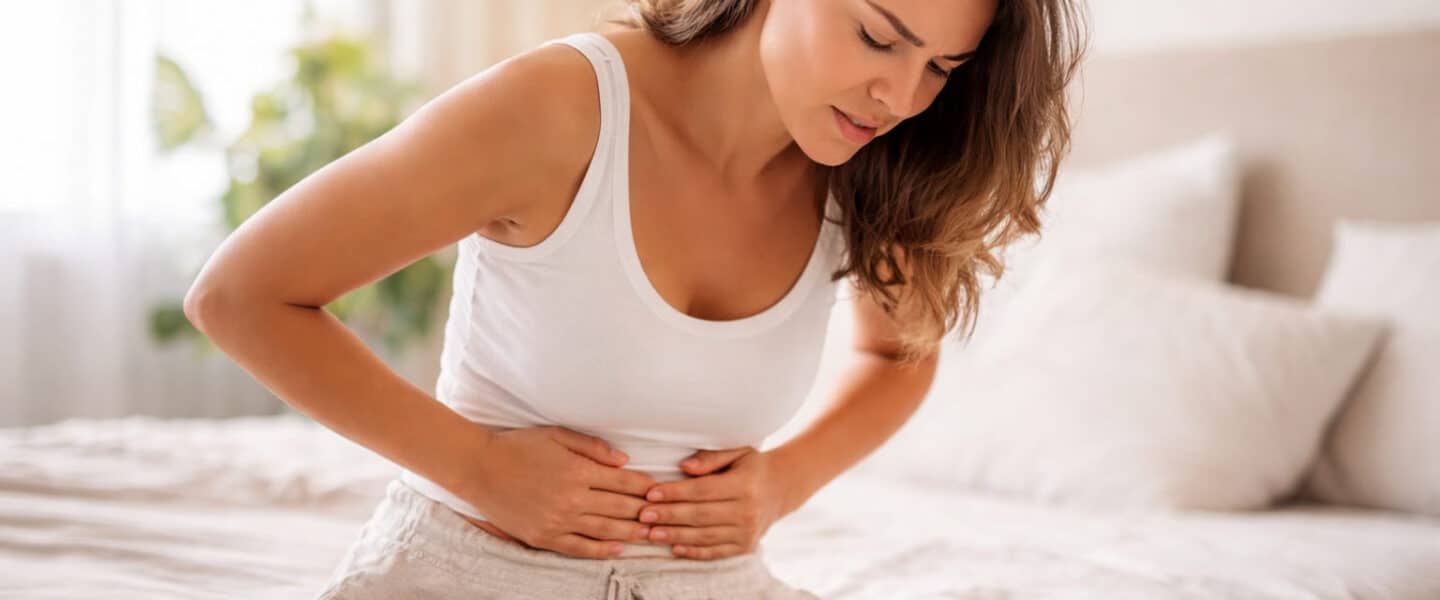 Vrouw zit op bed en houdt handen op haar buik ter hoogte van de navel vanwege buikpijn