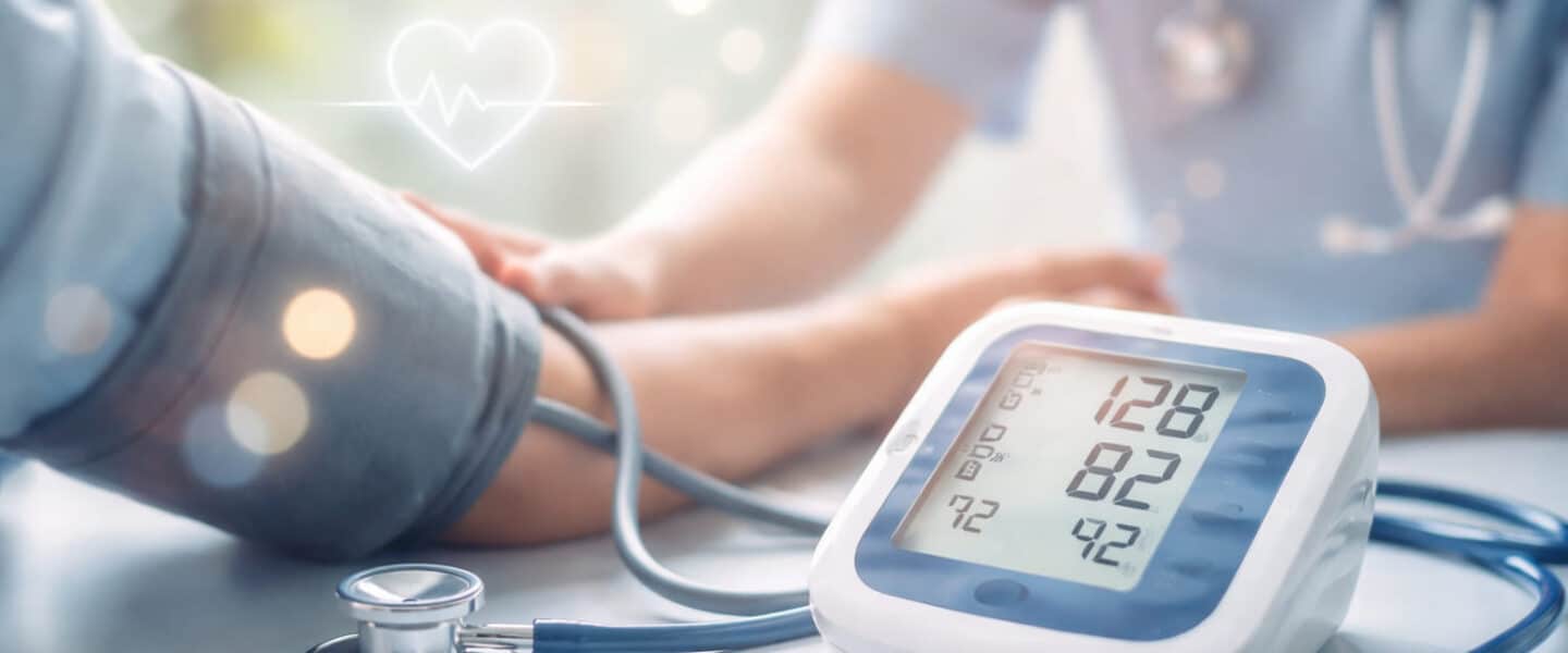 Wazige, breed uitgesneden headerafbeelding van een bloeddrukmeting in een rustige medische setting, met een digitale bloeddrukmeter op de voorgrond en een zorgverlener en patiënt zacht onscherp op de achtergrond.