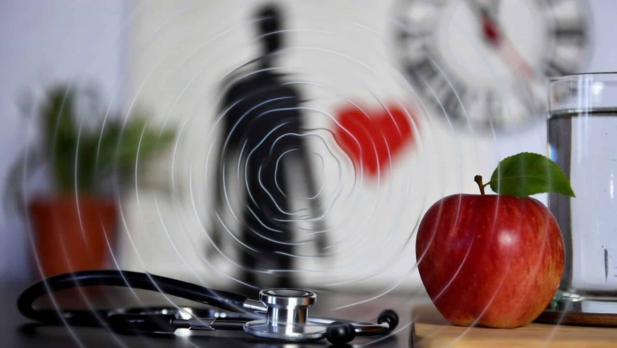Stethoscoop naast een rode appel en een glas water, met een vervaagd hart op de achtergrond als symbool voor bloeddruk en leefstijl