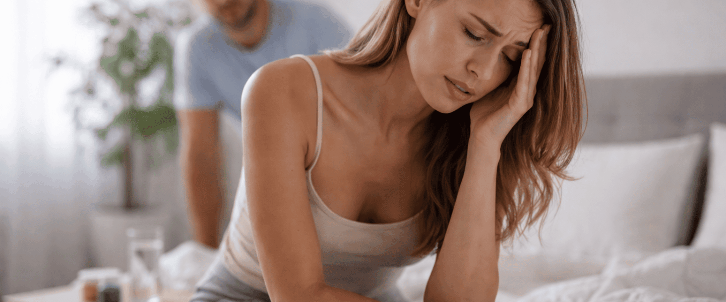 Jonge vrouw zit op de rand van een bed en houdt haar hoofd vast vanwege duizeligheid of een licht gevoel in het hoofd, terwijl op de achtergrond een bezorgde man nadert in een rustige, licht ingerichte slaapkamer.