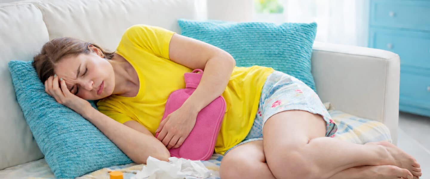 Vrouw ligt op de bank met PMS-klachten en houdt een roze kruik tegen haar onderbuik terwijl ze haar ogen gesloten houdt van de pijn.