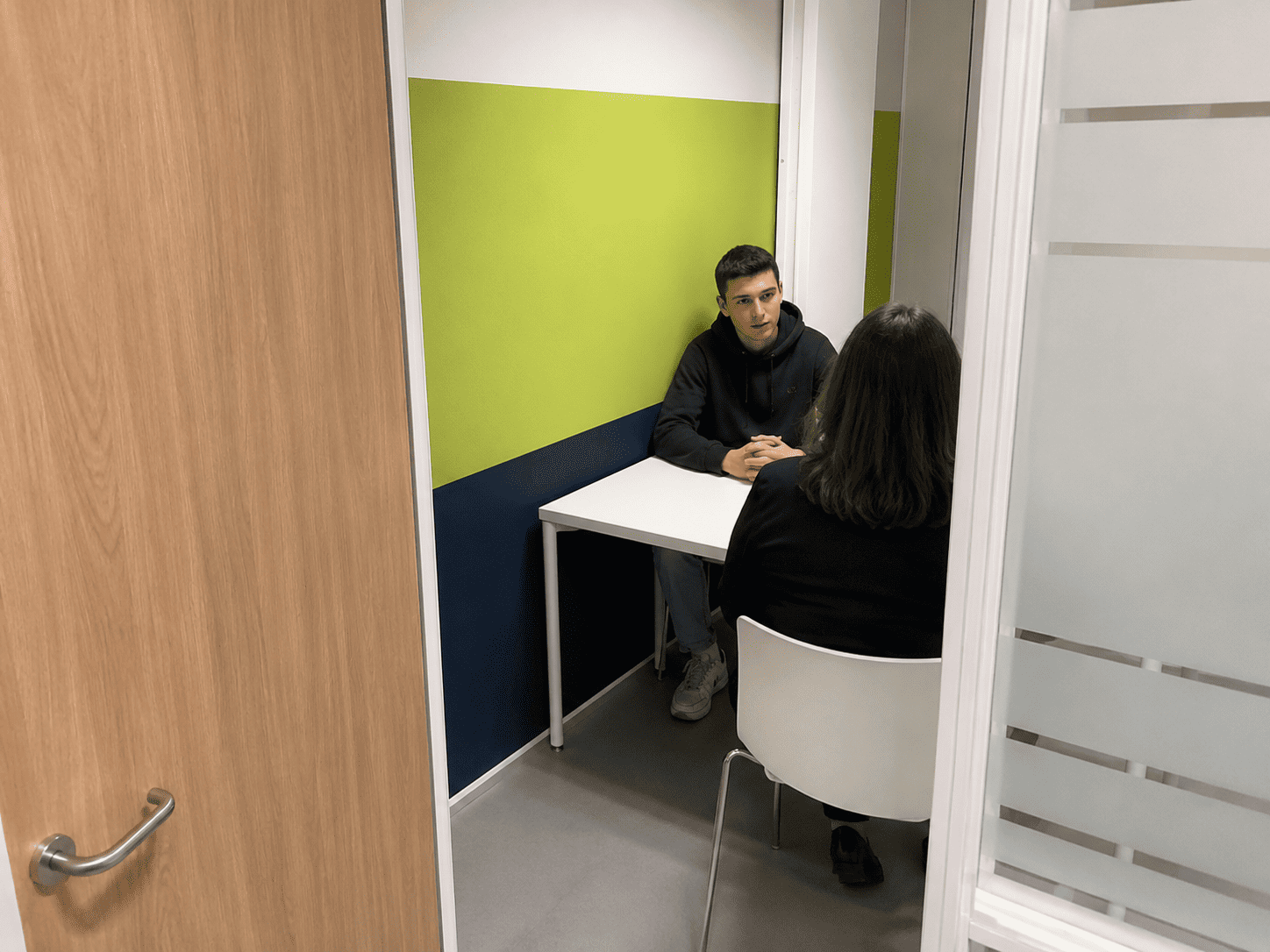 Cliënt in gesprek met een toezichthouder in een kleine spreekruimte van de reclassering, waarbij alleen de rug van de toezichthouder zichtbaar is.