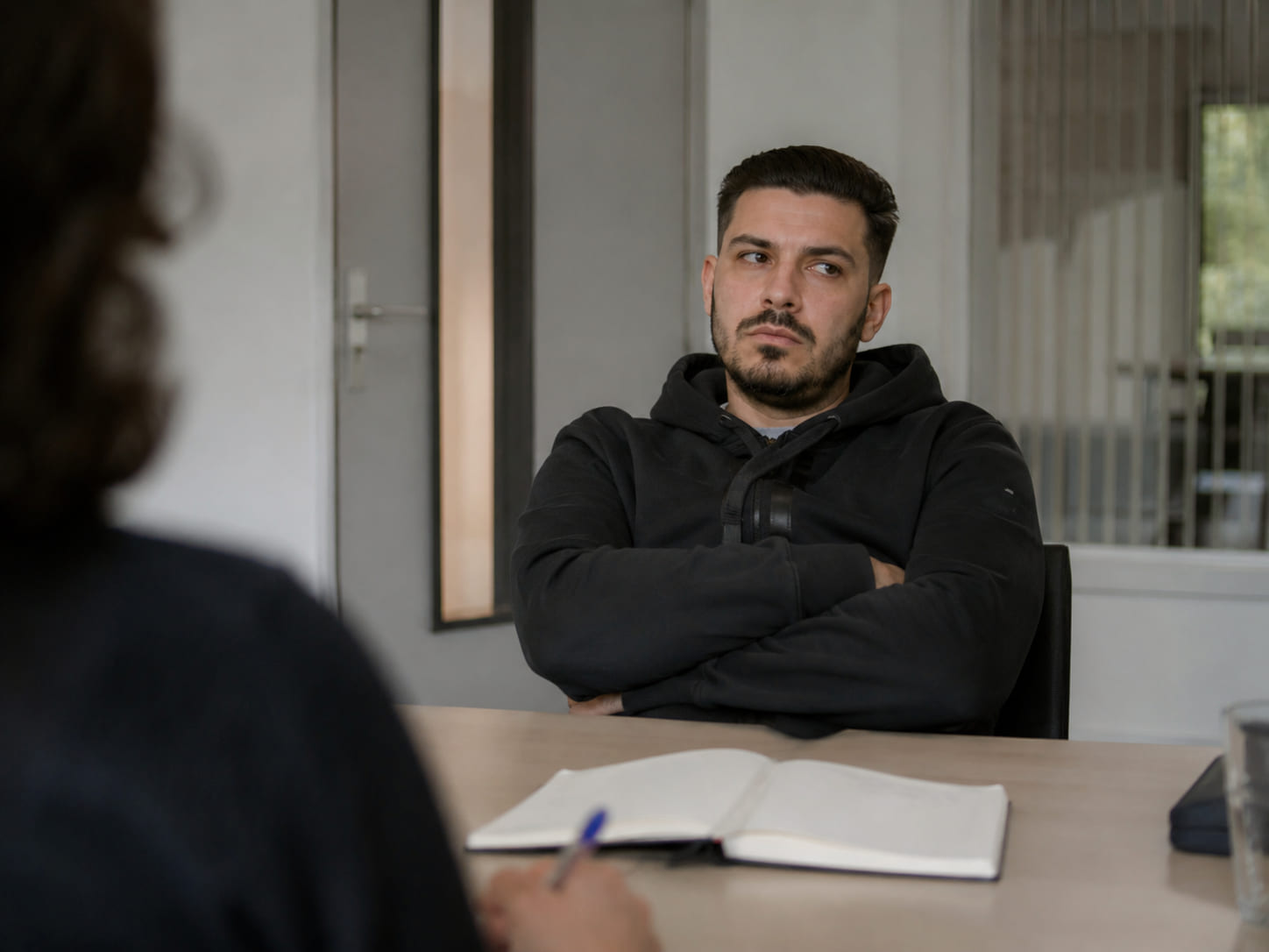 Cliënt zit met gekruiste armen en afgewende blik tegenover een toezichthouder tijdens een gesprek bij de reclassering.