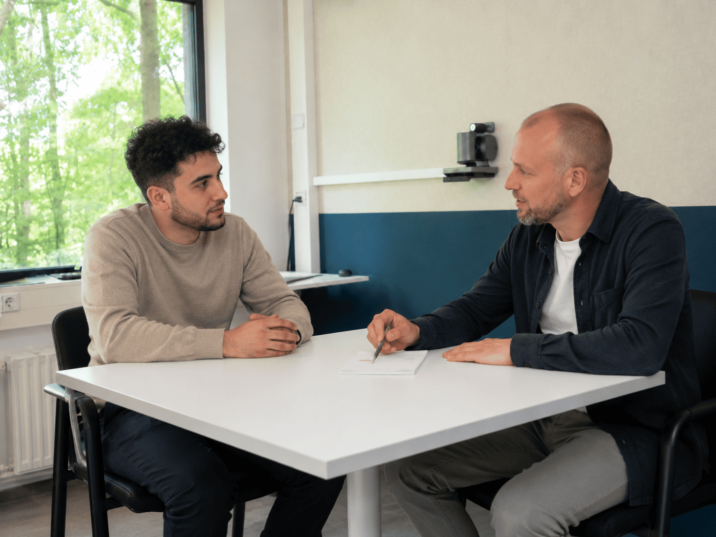 Cliënt en toezichthouder zitten aan een kleinere tafel in een lichte spreekkamer, in gesprek over begeleiding, keuzes en verantwoordelijkheid binnen het reclasseringstoezicht.