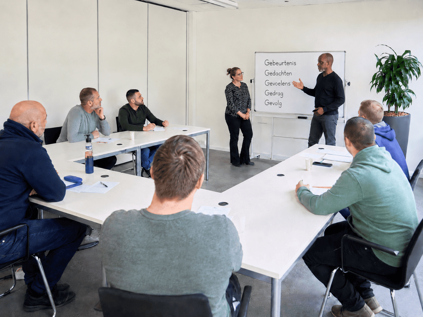 CoVa-training in een sobere reclasseringsruimte, met twee trainers bij een whiteboard waarop de 5 G’s staan en deelnemers die in U-vorm aan tafels zitten.