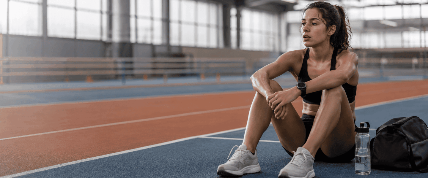 Vermoeide sporter zit na een training op een indoor atletiekbaan, passend bij RED-S en langdurig energietekort in de sport.