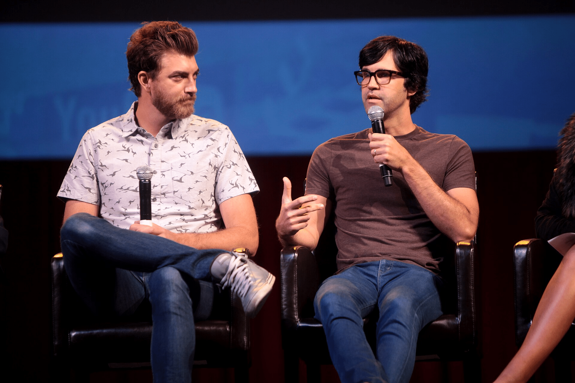 Twee mannen zitten op een podium tijdens een panelgesprek en houden microfoons vast; de ene kijkt opzij, terwijl de ander spreekt tegen het publiek.
