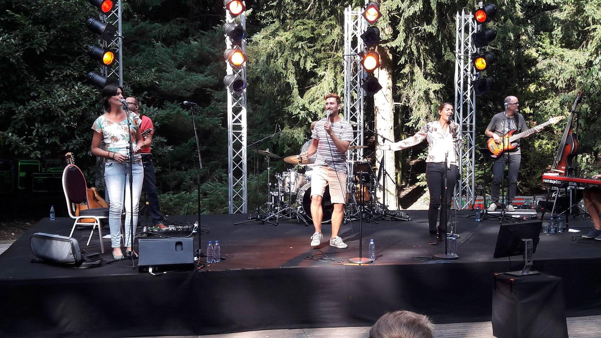 De christelijke band Sela treedt op tijdens een openluchtconcert, met zangers, muzikanten, microfoons en podiumverlichting tussen de bomen.