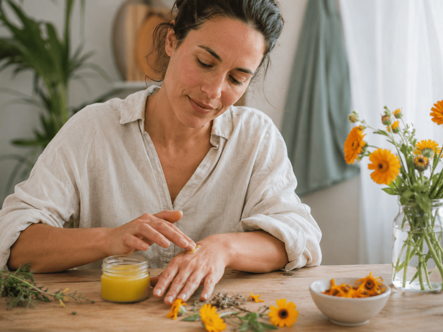Vrouw brengt calendulazalf aan op haar hand, met goudsbloemen en gedroogde bloemblaadjes op tafel.