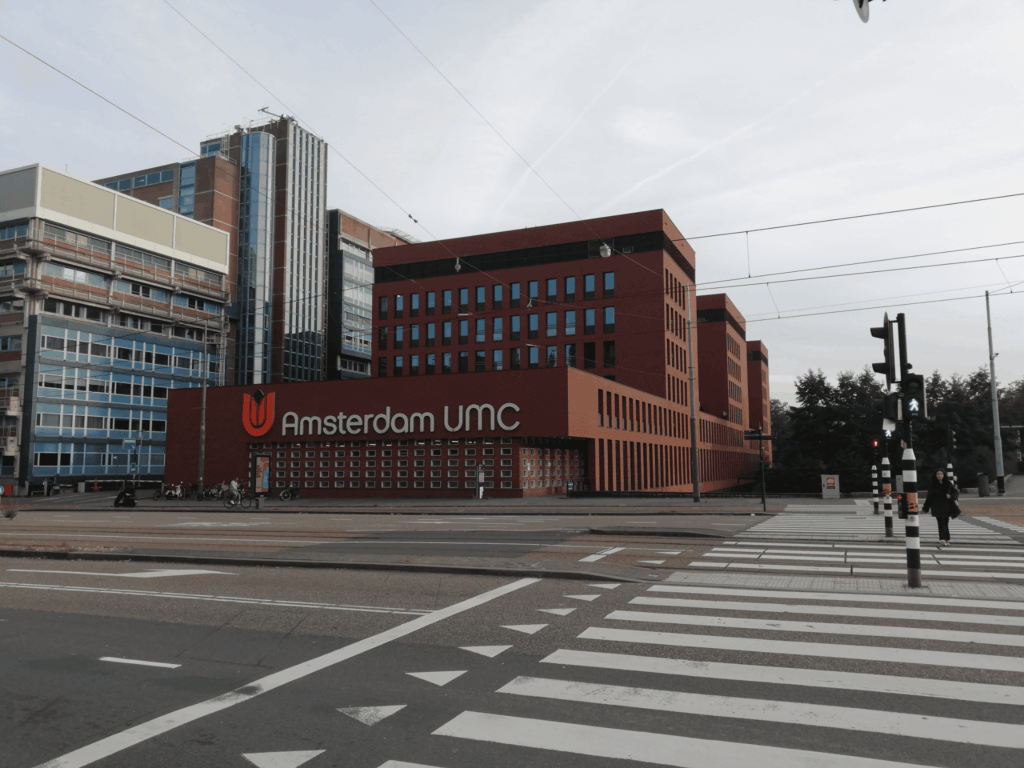 Buitenzijde van Amsterdam UMC met rode bakstenen gevel, ziekenhuisgebouwen, tramdraden en zebrapad op de voorgrond.