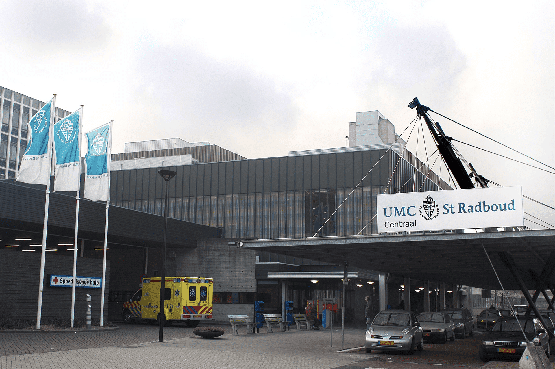 Ingang van het UMC St Radboud in Nijmegen met vlaggen, ambulance, geparkeerde auto’s en het bord “UMC St Radboud Centraal”.